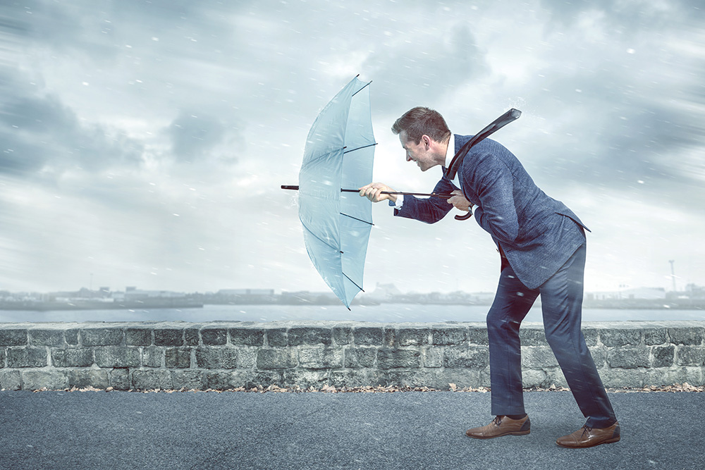 Businessman with an umbrella standing firm against a powerful headwind, epitomizing resilience in the face of challenges.